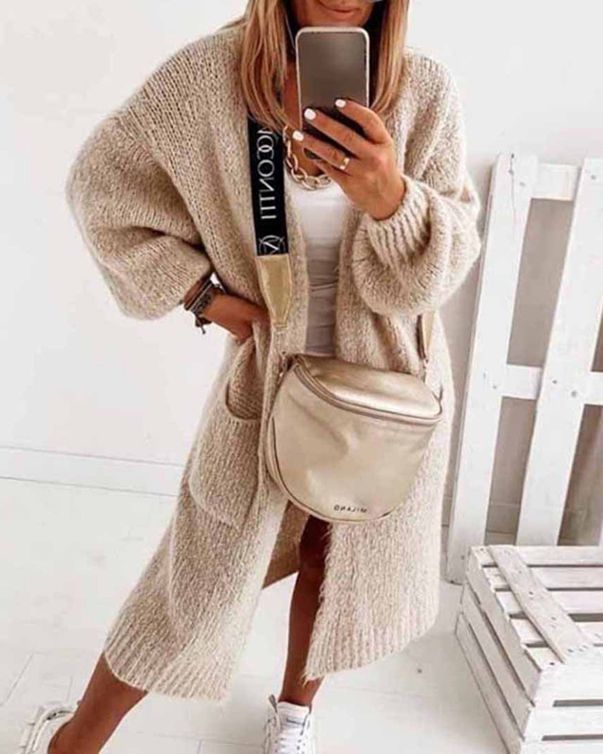 Frau in beigem Strickmantel mit Umhängetasche, weißem Top und Sneakers, macht Selfie. Modetrend, lässiger Herbstlook, Damenmode.