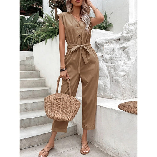 Frau in braunem Jumpsuit mit Gürtel, hält geflochtene Korbtasche, steht auf Treppe vor weißer Wand, Sommermode, lässiger Stil.
