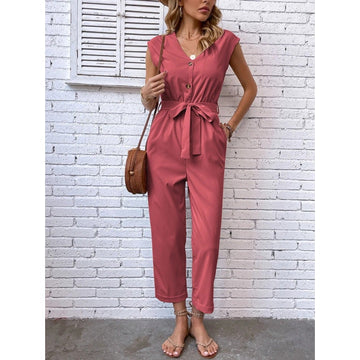 Frau in rosa Jumpsuit vor weißer Backsteinwand, trägt braune Umhängetasche und Sandalen. Sommermode, lässiger Stil, trendiges Outfit.