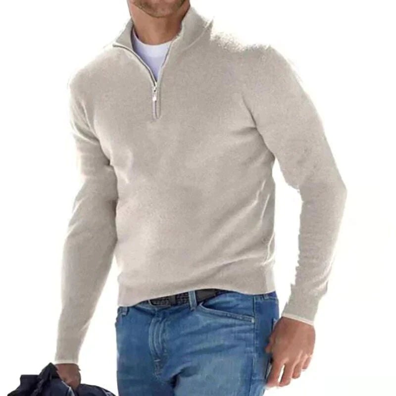 Mann in beigem Pullover mit Reißverschluss und blauer Jeans, hält eine Jacke. Modischer Freizeitlook für Herren, ideal für kühle Tage.