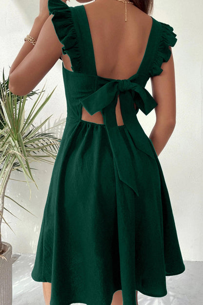 Frau in dunkelgrünem Sommerkleid mit Rückenausschnitt und Schleife, neben einer Zimmerpflanze. Elegantes, ärmelloses Design, ideal für warme Tage.