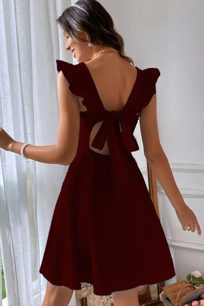Frau in elegantem, ärmellosem, weinrotem Kleid mit Rückenausschnitt und Schleife, steht vor einem Fenster. Perfekt für Sommermode und Abendveranstaltungen.