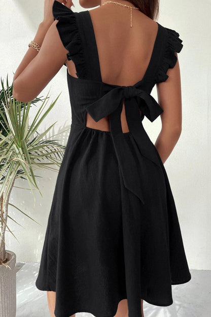 Frau in schwarzem Sommerkleid mit Rückenausschnitt und Schleife, neben Zimmerpflanze. Elegantes, ärmelloses Design, ideal für warme Tage.