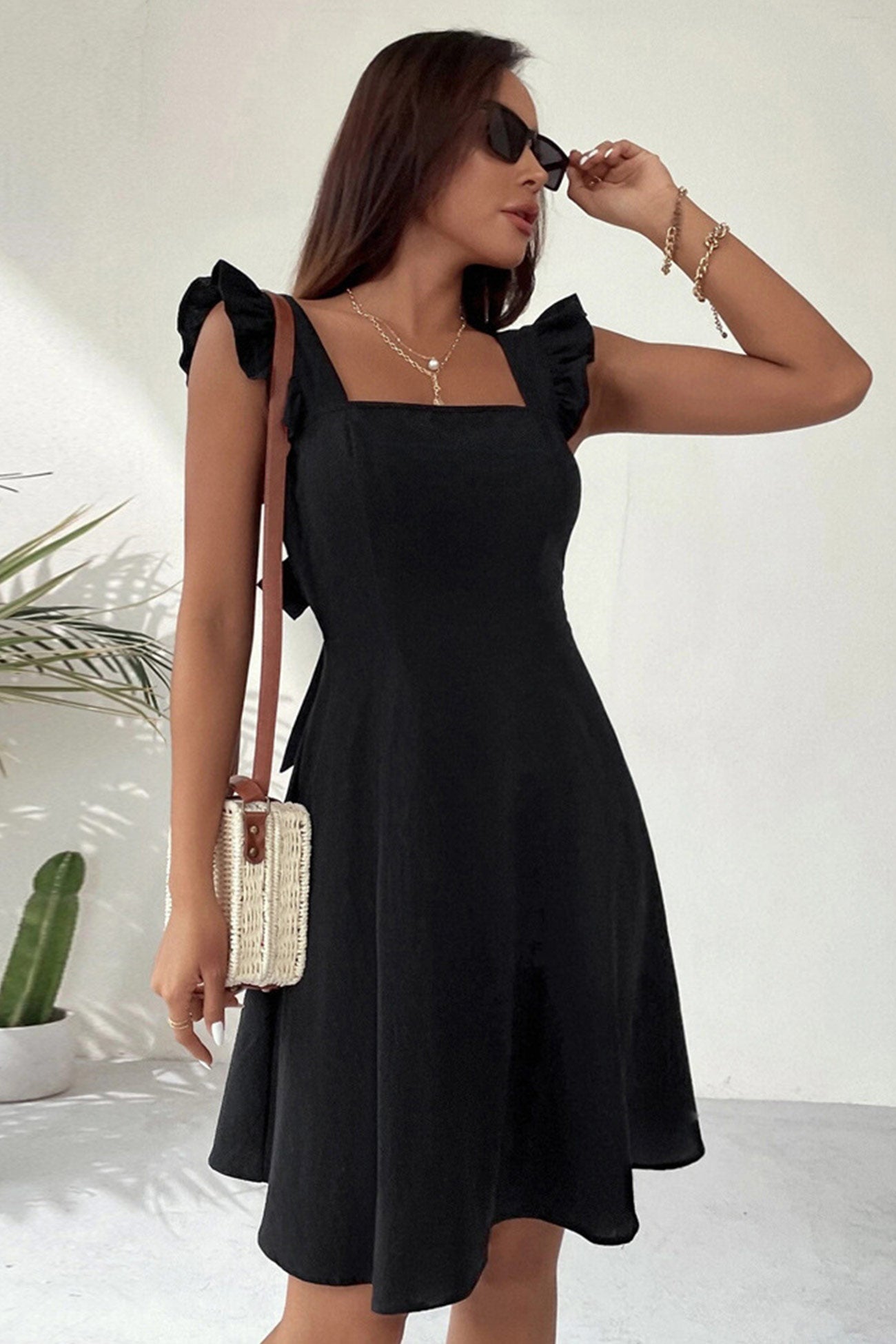 Frau in schwarzem Sommerkleid mit Rüschenärmeln, Sonnenbrille und Strohtasche, steht vor weißem Hintergrund mit Pflanze. Elegante Mode, Sommeroutfit.