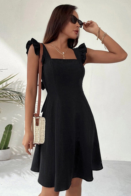 Frau in schwarzem Sommerkleid mit Rüschenärmeln, Sonnenbrille und Strohtasche, steht vor weißem Hintergrund mit Pflanze. Elegante Mode, Sommeroutfit.
