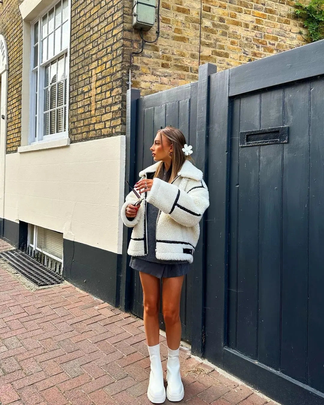 Frau in modischer weißer Teddyjacke und schwarzen Minirock, steht vor einer Backsteinmauer, hält ein Handy, trägt weiße Stiefel. Streetstyle, Herbstmode.