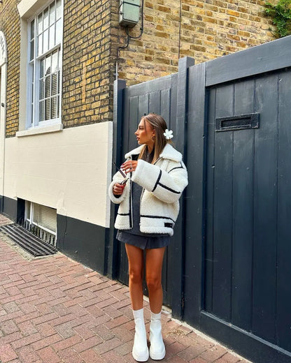 Frau in modischer weißer Teddyjacke und schwarzen Minirock, steht vor einer Backsteinmauer, hält ein Handy, trägt weiße Stiefel. Streetstyle, Herbstmode.