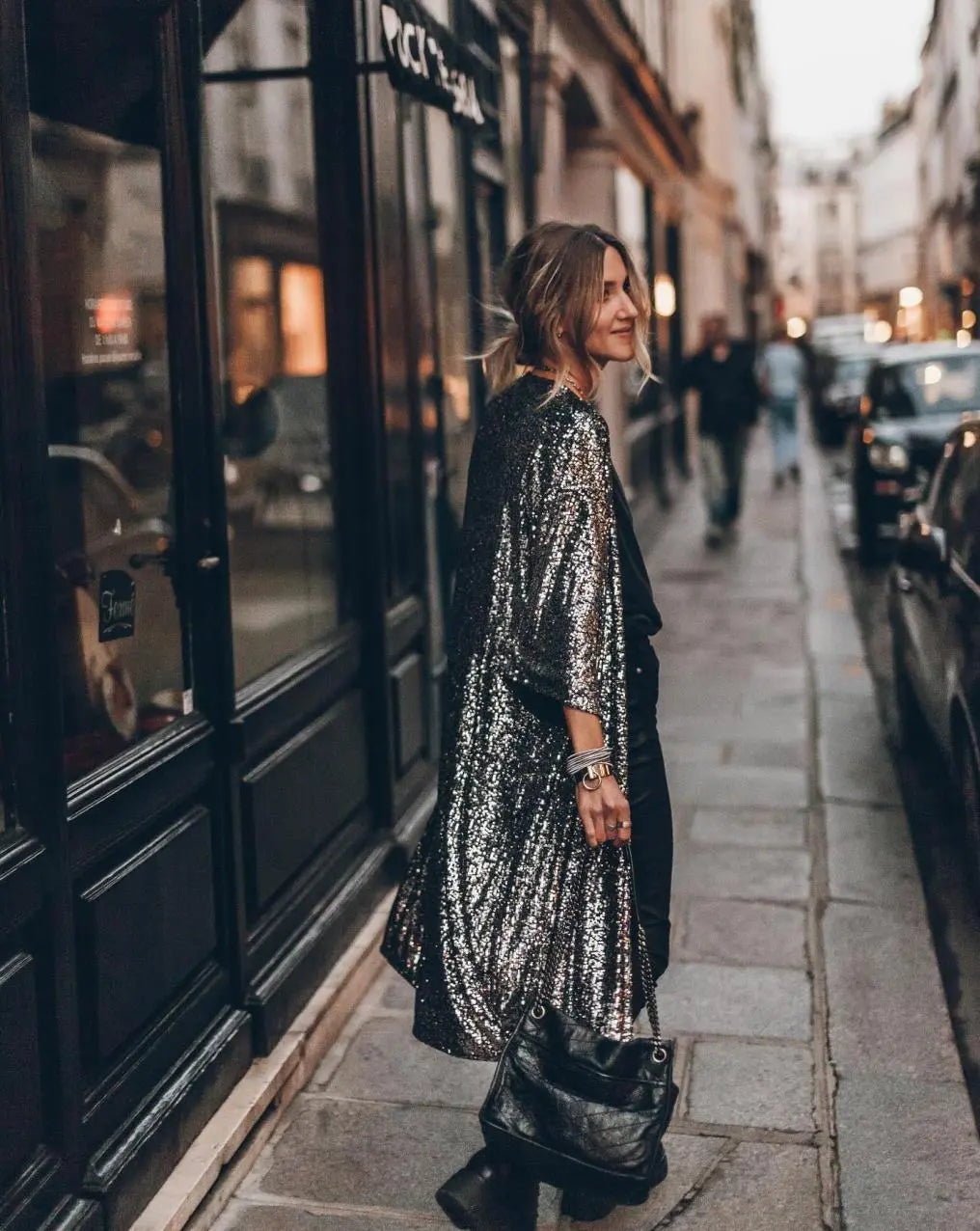 Frau in glitzerndem silbernen Mantel auf Pariser Straße, trägt schwarze Tasche. Abendstimmung, modisch, urbaner Stil.