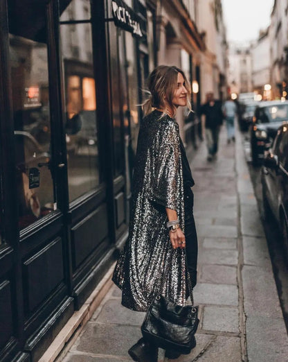 Frau in glitzerndem silbernen Mantel auf Pariser Straße, trägt schwarze Tasche. Abendstimmung, modisch, urbaner Stil.