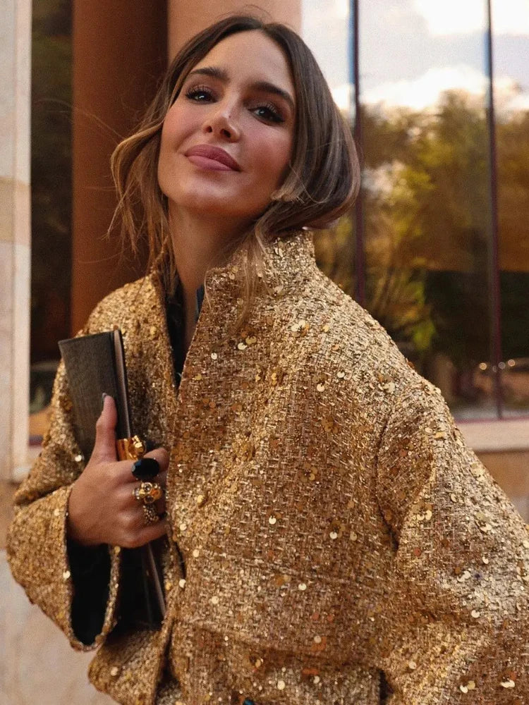 Frau in goldener Paillettenjacke hält schwarze Clutch, lächelt in die Kamera. Elegante Mode, glamouröser Look, stilvolle Abendgarderobe.