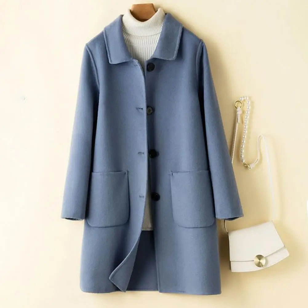 Hellblauer Damenmantel mit Knöpfen und Taschen, kombiniert mit weißem Rollkragenpullover und kleiner weißer Handtasche an cremefarbener Wand.