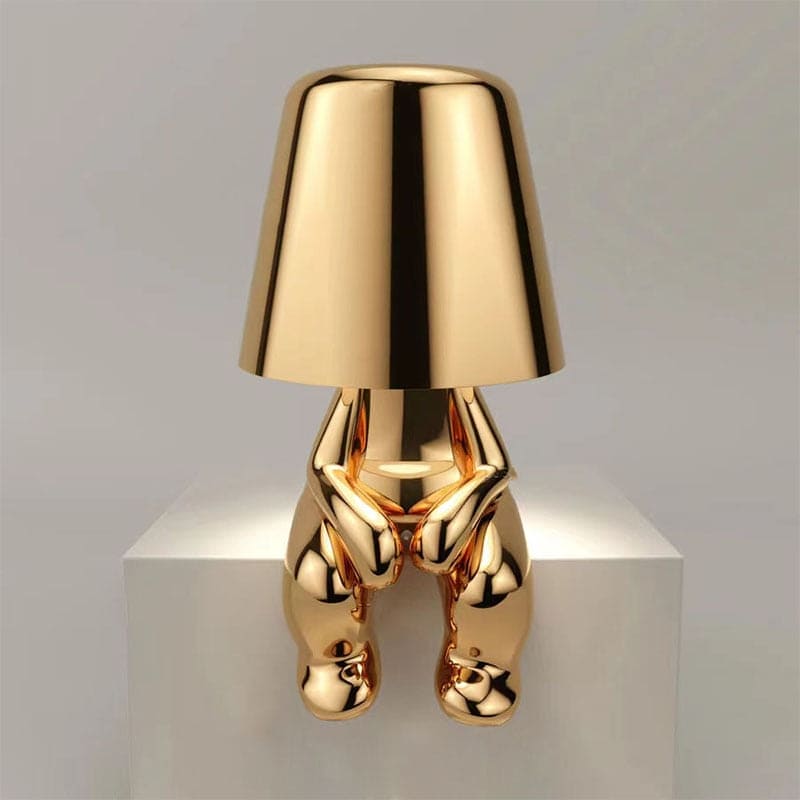 Goldene Designer-Tischlampe in Form einer sitzenden Figur auf weißem Podest. Modernes, elegantes Wohnaccessoire, ideal für stilvolle Inneneinrichtung.