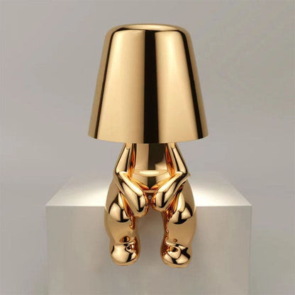 Goldene Designer-Tischlampe in Form einer sitzenden Figur auf weißem Podest. Modernes, elegantes Wohnaccessoire, ideal für stilvolle Inneneinrichtung.