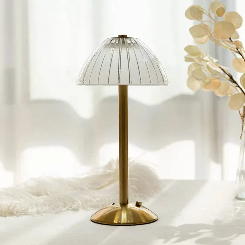 Goldene Tischlampe mit transparentem Glasschirm auf weißem Tisch, im Hintergrund helle Vorhänge und dekorative Trockenblumen. Ideal für modernes Wohndesign.