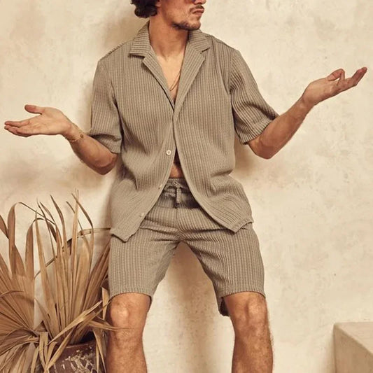 Mann in beige geripptem Kurzarmhemd und Shorts, sitzt lässig vor neutralem Hintergrund, moderne Herrenmode, entspannter Sommerlook.