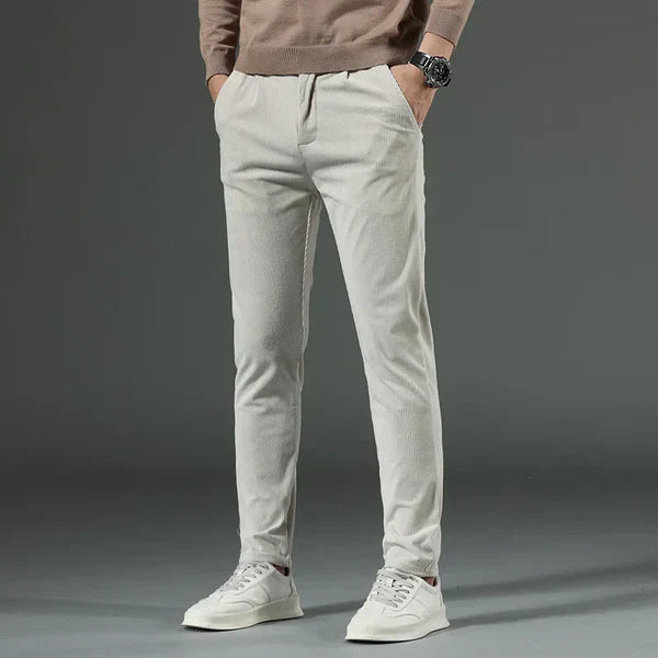 Mann in grauer Chinohose und weißen Sneakers, brauner Pullover, Hände in den Taschen. Modische Herrenbekleidung, lässiger Stil.