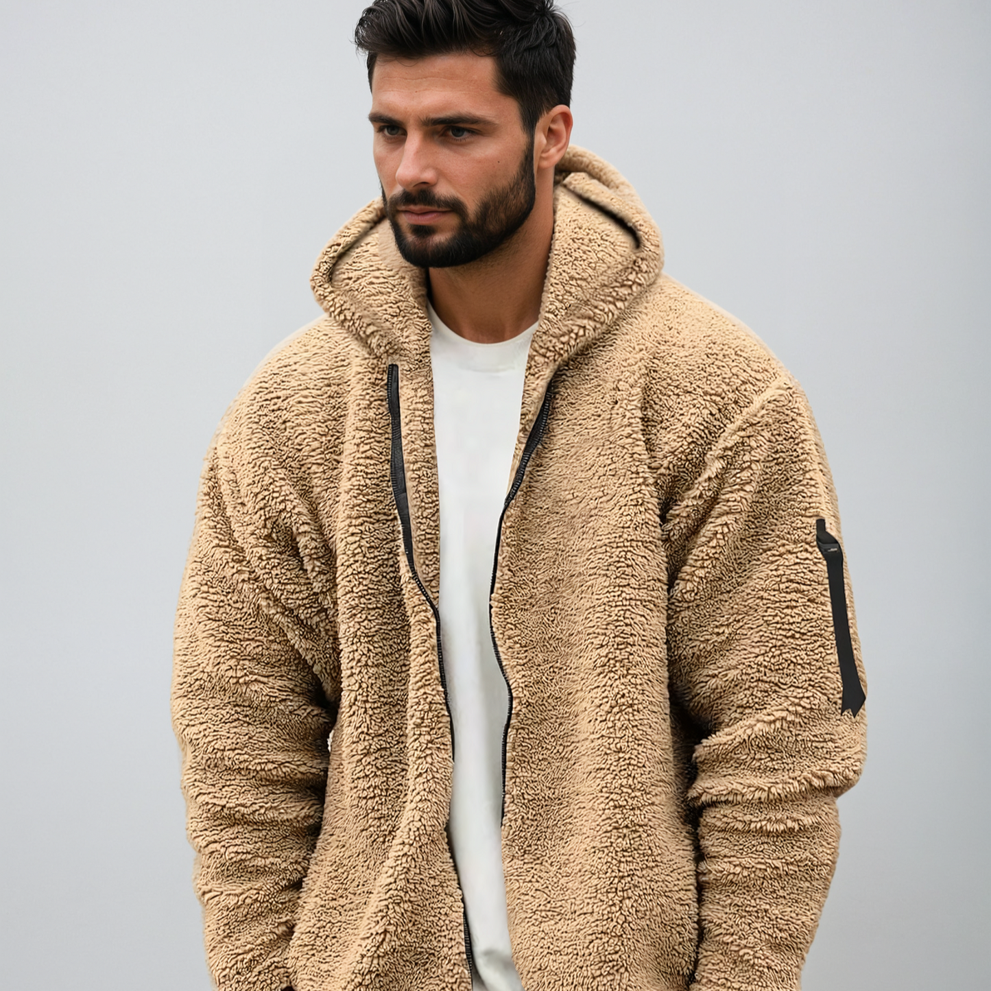 Mann in beiger Teddyfleece-Jacke mit Kapuze, Reißverschluss und Tasche, vor grauem Hintergrund. Modetrend, Herrenbekleidung, Winterjacke.