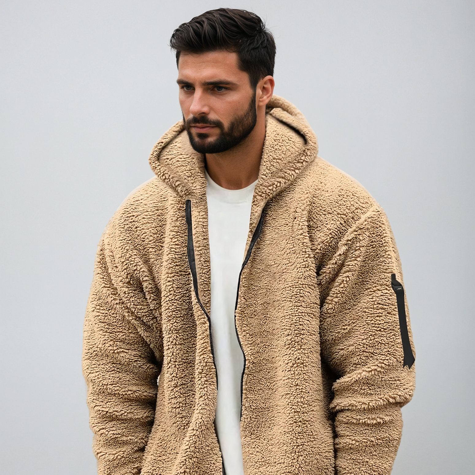 Mann in beiger Teddyfleece-Jacke mit Kapuze, Reißverschluss und Armtasche vor grauem Hintergrund. Modetrend, Herrenbekleidung, Winterjacke.