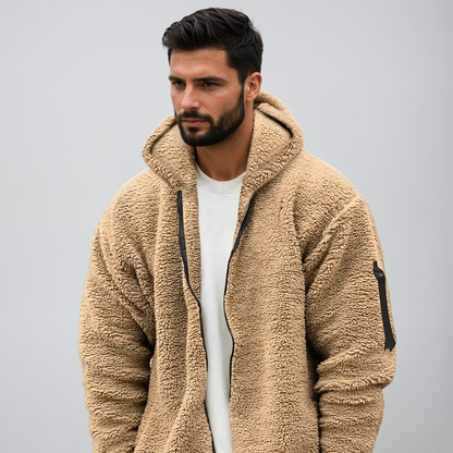 Mann in beiger Teddyfleece-Jacke mit Kapuze, Reißverschluss und Armtasche vor grauem Hintergrund. Modetrend, Herrenbekleidung, Winterjacke.