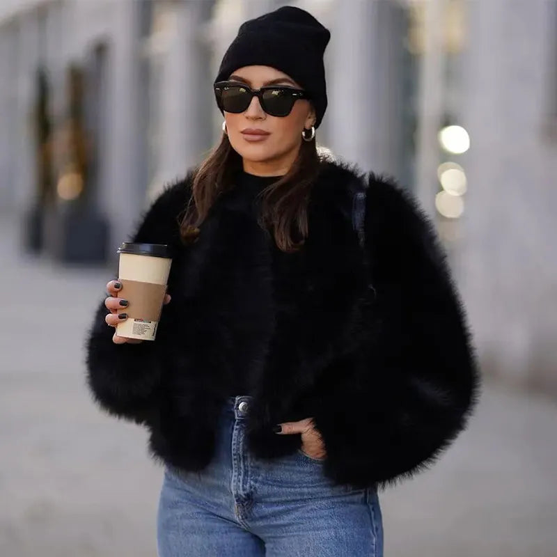 Frau in schwarzer Felljacke, blauer Jeans und Mütze hält Kaffeebecher. Modetrend, Winteroutfit, Streetstyle, Sonnenbrille, urbaner Look.