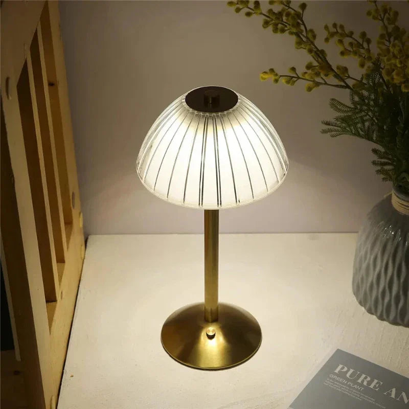 Goldene Tischlampe mit geriffeltem Glasschirm auf Holztisch, neben Vase mit Blumen und Buch. Elegante Beleuchtung, modernes Design.