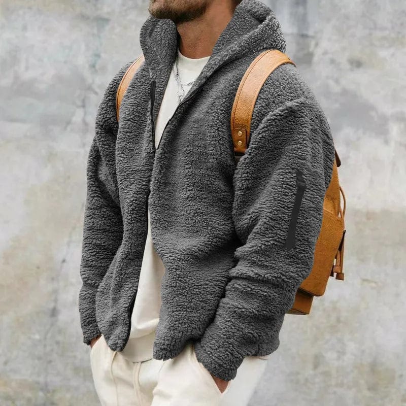 Mann in grauer Fleecejacke mit braunem Rucksack vor grauer Wand. Modischer Winterlook, warme Kleidung, Herrenmode, Outdoor-Stil.