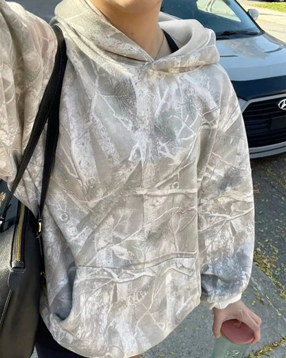 Person in einem grauen Camouflage-Hoodie mit Blättermuster, hält einen rosa Getränkebecher. Im Hintergrund ein geparktes Auto auf der Straße.