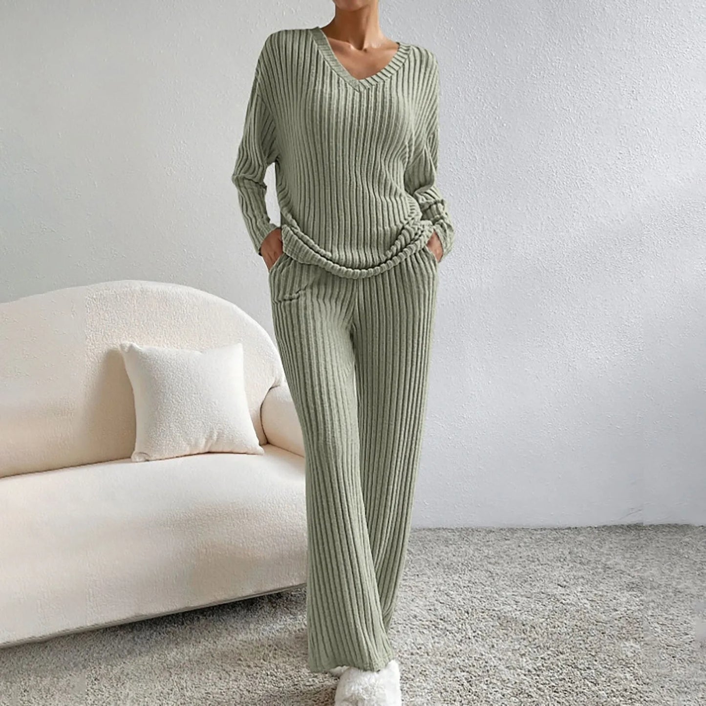 Frau in grünem Strick-Loungewear-Set, bestehend aus Pullover und Hose, steht in modernem Wohnzimmer mit beigem Sofa und Kissen.
