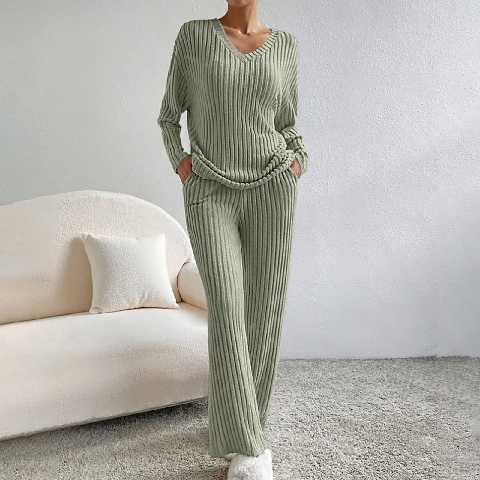 Frau in grünem Strick-Loungewear-Set, bestehend aus Pullover und Hose, steht in modernem Wohnzimmer mit beigem Sofa und Kissen.