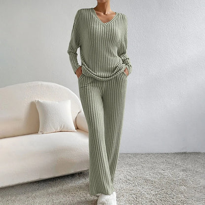Frau in grünem Strick-Loungewear-Set, bestehend aus Pullover und Hose, steht in modernem Wohnzimmer mit beigem Sofa und Kissen.