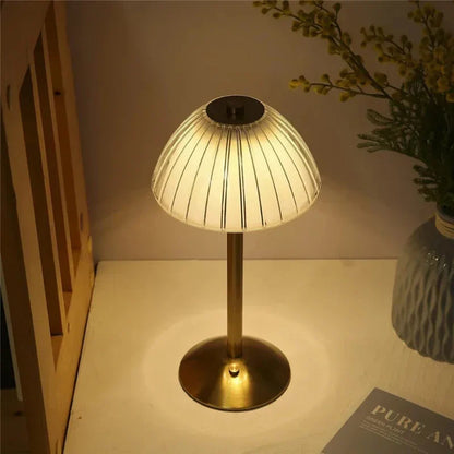 Goldene Tischlampe mit geriffeltem Schirm auf Holzoberfläche, neben Vase mit Blumen und Buch. Elegante Beleuchtung, modernes Design.