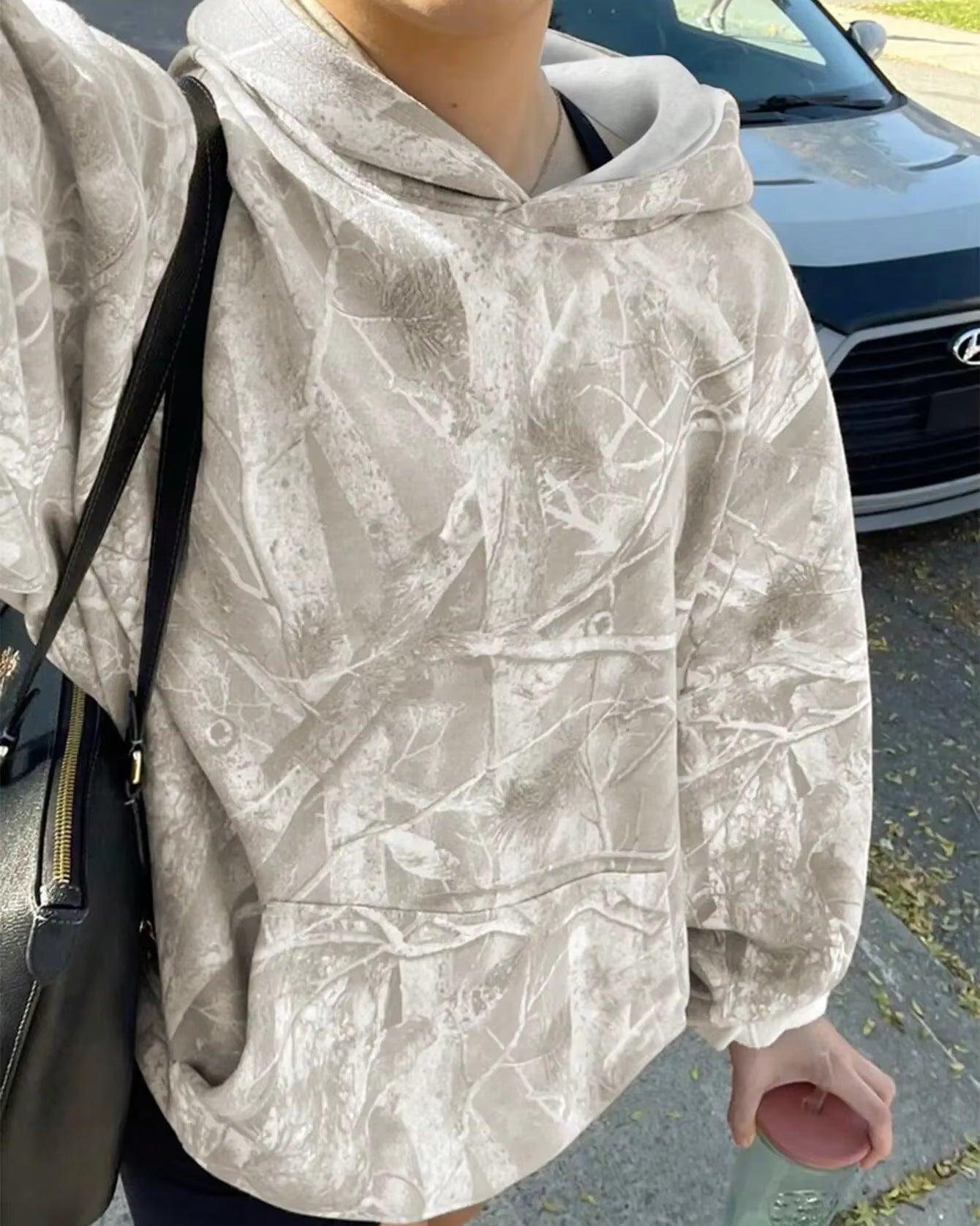 Person in grauem Camouflage-Hoodie mit schwarzer Tasche und Wasserflasche, steht vor einem geparkten Hyundai-Auto auf der Straße.