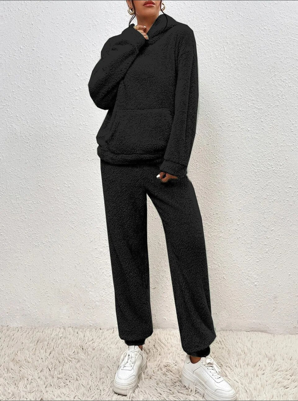 Frau in schwarzem Fleece-Hoodie und Jogginghose, weiße Sneaker, steht vor weißer Wand auf Teppich. Bequeme Freizeitmode, lässiger Look.