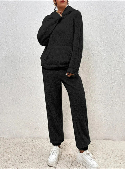 Frau in schwarzem Fleece-Hoodie und Jogginghose, weiße Sneaker, steht vor weißer Wand auf Teppich. Bequeme Freizeitmode, lässiger Look.