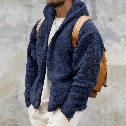 Mann in dunkelblauer Fleecejacke mit Kapuze und braunem Rucksack vor grauer Wand. Modetrend, Herrenbekleidung, Herbstmode.