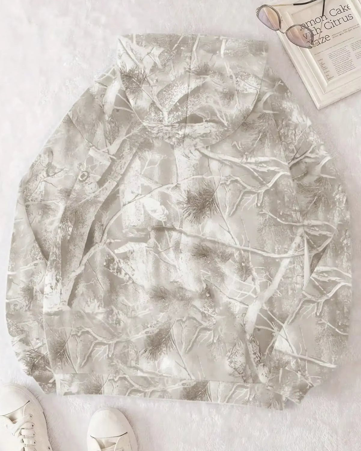 Tarnmuster Hoodie auf weißem Hintergrund, neben Sonnenbrille und Buch. Perfekt für Outdoor-Aktivitäten, Jagd und Freizeitmode.