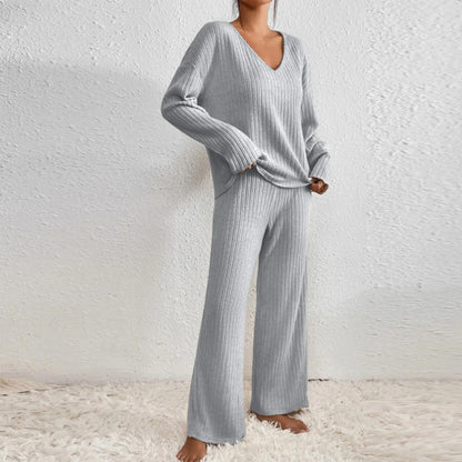 Frau in grauem Strick-Loungewear-Set, bestehend aus V-Pullover und Hose, steht barfuß auf Teppich vor weißer Wand. Gemütliche Mode, Homewear.