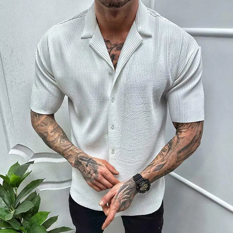 Mann in weißem, kurzärmligem Hemd mit Tätowierungen auf den Armen, steht vor einer weißen Wand. Mode, Herrenbekleidung, Sommerlook.