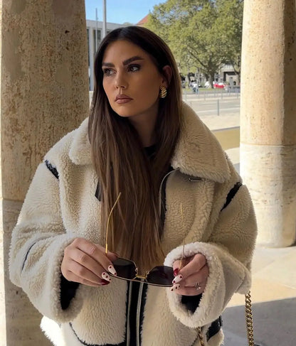 Frau in weißem Teddy-Mantel hält Sonnenbrille, steht vor Säulen. Herbstmode, stilvoll, urbaner Hintergrund. Elegante, lange braune Haare.