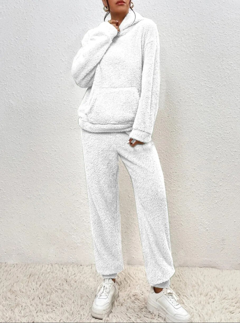 Weiße Fleece-Loungewear-Set mit Kapuzenpullover und Jogginghose, getragen von einer Person vor einer strukturierten Wand. Bequeme Freizeitmode.