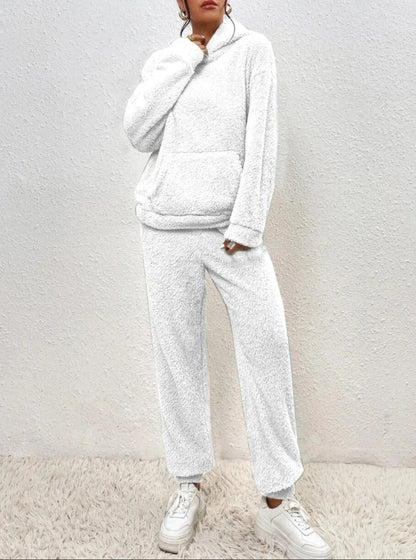 Weiße Fleece-Loungewear-Set mit Kapuzenpullover und Jogginghose, getragen von einer Person vor einer strukturierten Wand. Bequeme Freizeitmode.