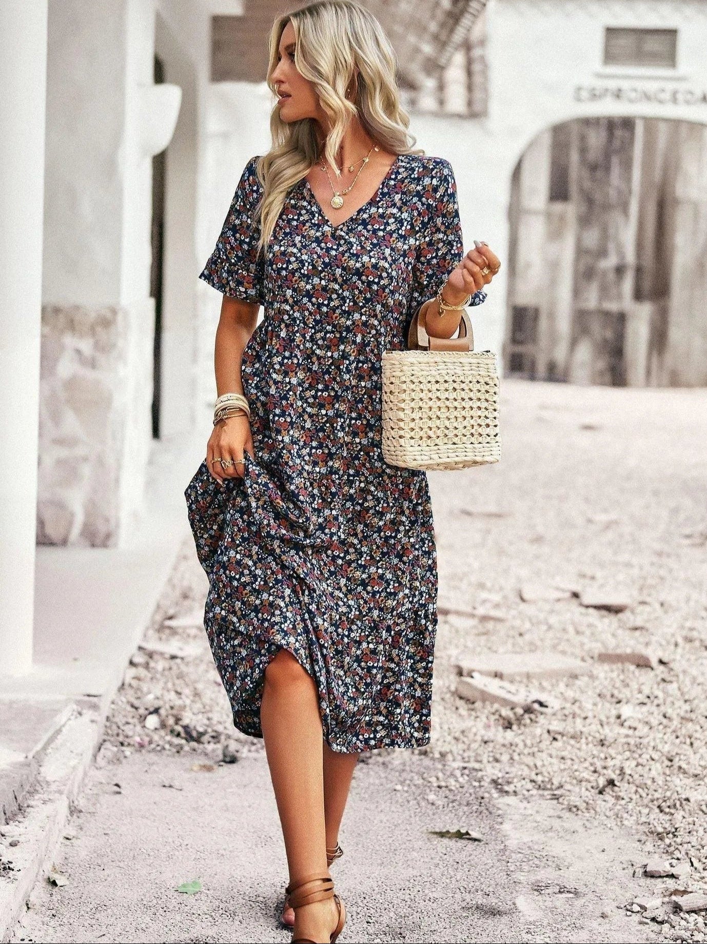 Blonde Frau in geblümtem Sommerkleid, trägt Strohtasche, spaziert auf gepflastertem Weg. Modetrend, Sommermode, Damenkleid, Freizeitlook.