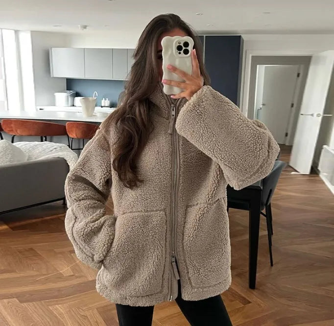 Frau in beiger Teddyjacke macht Selfie in modernem Wohnzimmer; gemütliche Wintermode, trendige Damenbekleidung, stilvolle Inneneinrichtung.