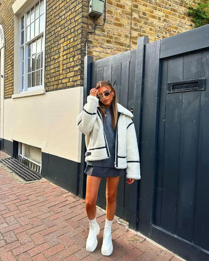 Frau in weißer Teddyjacke und grauem Kleid, trägt Sonnenbrille und weiße Stiefel, steht vor einer Backsteinmauer und Holztür. Streetstyle, Mode.