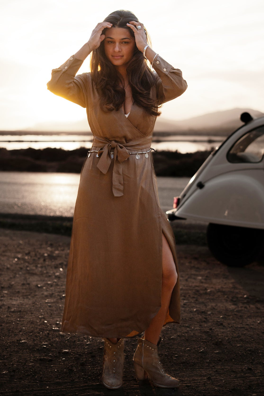 Frau in langem, braunem Kleid posiert vor einem Oldtimer bei Sonnenuntergang. Modetrend, Vintage-Auto, Abendstimmung, stilvolle Kleidung.