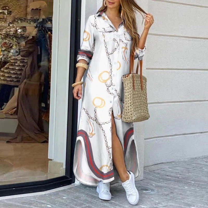 Frau in weißem Maxikleid mit Kettenmuster, trägt Strohtasche und weiße Sneaker, steht vor einem Schaufenster. Elegante Sommermode, lässiger Stil.