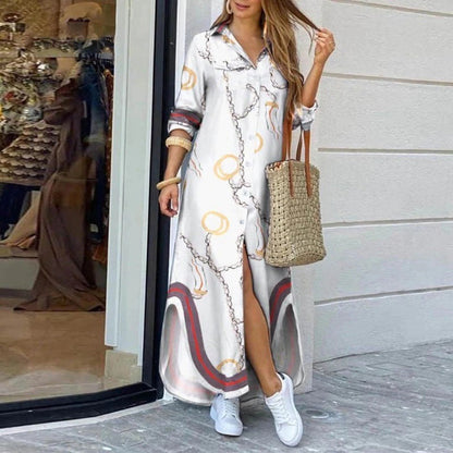 Frau in weißem Maxikleid mit Kettenmuster, trägt Strohtasche und weiße Sneaker, steht vor einem Schaufenster. Elegante Sommermode, lässiger Stil.