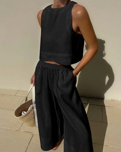 Frau in schwarzem Leinen-Zweiteiler mit ärmellosem Top und weiter Hose, hält Strohtasche. Sommermode, minimalistischer Stil, elegantes Outfit.