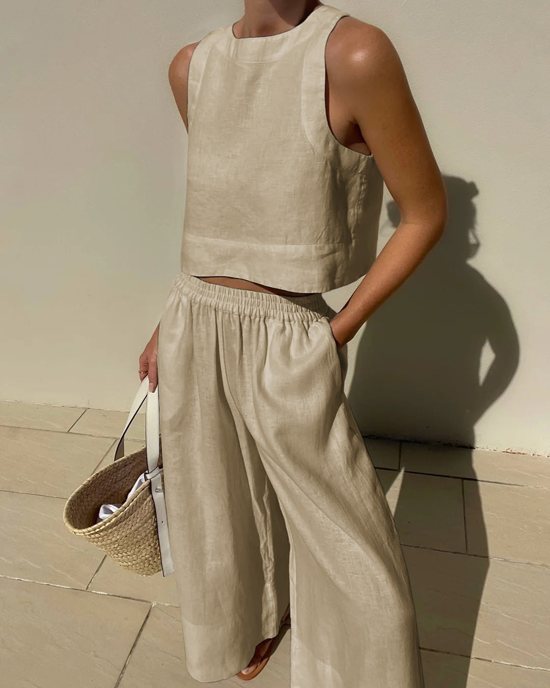 Frau in beigem Leinen-Zweiteiler mit ärmellosem Top und weiter Hose, hält Strohtasche. Sommermode, minimalistischer Stil, elegantes Design.