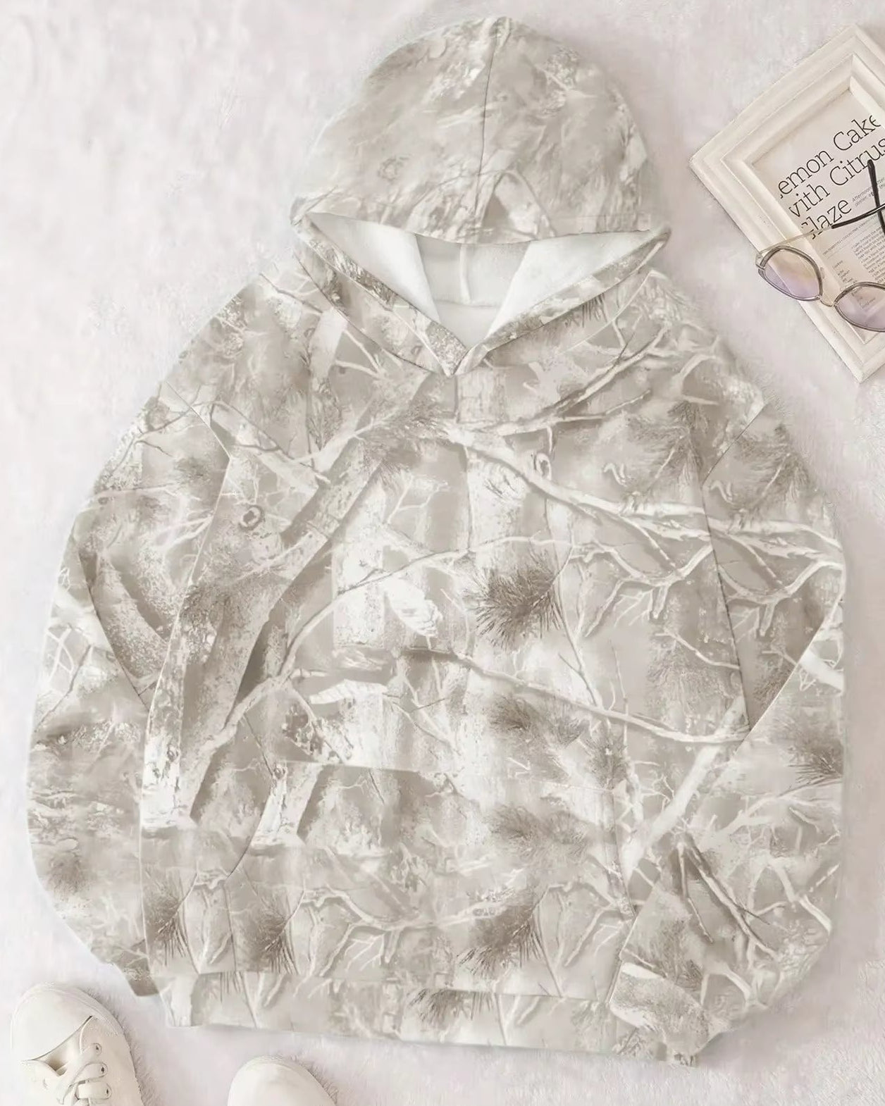 Grauer Camouflage-Hoodie mit Waldmuster auf weißem Hintergrund, neben einer Brille und einem Buch. Bequeme Freizeitmode für Outdoor-Aktivitäten.